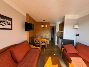 Een knus ingerichte kamer met een houten wand, een aan de muur bevestigde televisie, een eettafel met stoelen, een kitchenette en een bank met rode kussens.