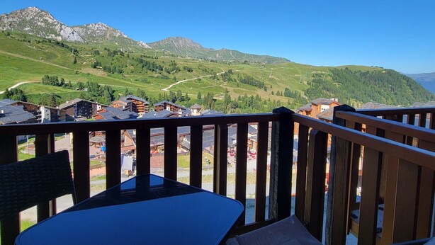 A balcony with a view of a mountainous landscape, wooden railings, and a table with a chair.