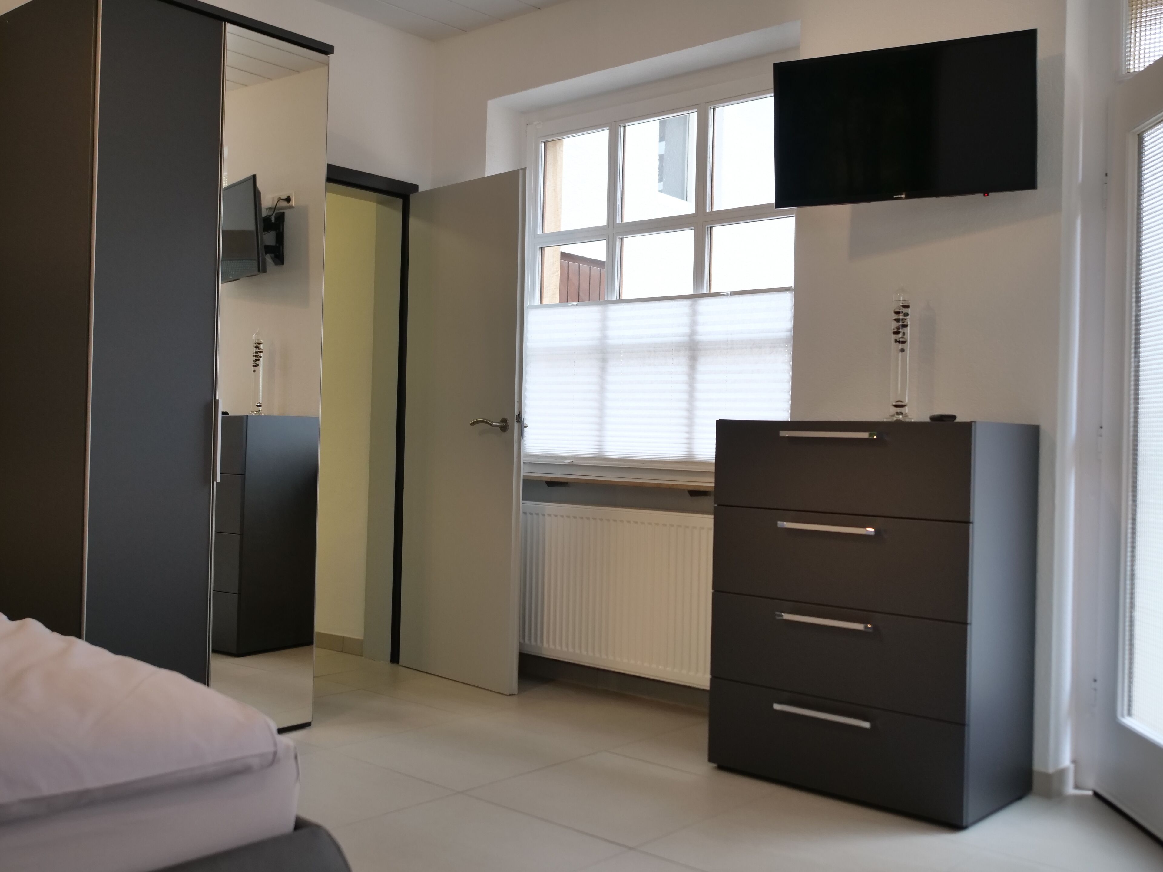 A bedroom with a bed, a wardrobe, a dresser, a window with blinds, and a mounted TV.