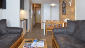 A modern living room with two grey sofas, a wooden coffee table with magazines, a TV mounted on the wall, and a dining area with a wooden table and chairs. - Renovated apartment at slopes with balcony (Belle Plagne)