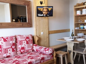 A cozy room with a wooden sofa, a television mounted on the wall, a wooden table with dishes, and a mirror above the sofa.