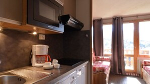 Et kompakt køkken med mikroovn, kaffemaskine og vask. Et lille spiseområde med et bord og stole kan ses gennem en åben dør.
