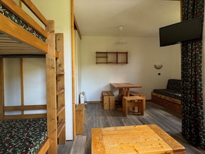Una habitación compacta con una cama nido, una pequeña mesa de madera, un sofá y un televisor fijado en la pared.