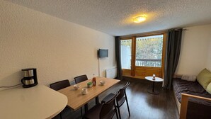 Una habitación acogedora con una mesa de comedor de madera, sillas, una cafetera, un televisor y un sofá con vista a los árboles a través de un ventanal grande.
