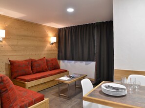 A cozy room with a wooden wall, a red sofa, a small table, and a dining area with chairs and plates.