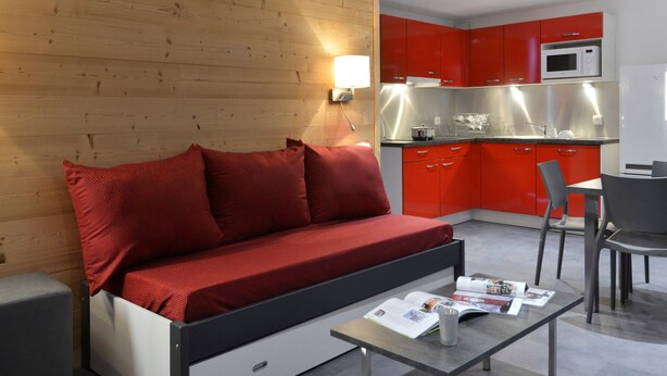 Ein moderner Wohnraum mit einem roten Sofa, einem kleinen Esstisch und einer Küchenzeile mit roten Schränken und Edelstahlgeräten.
