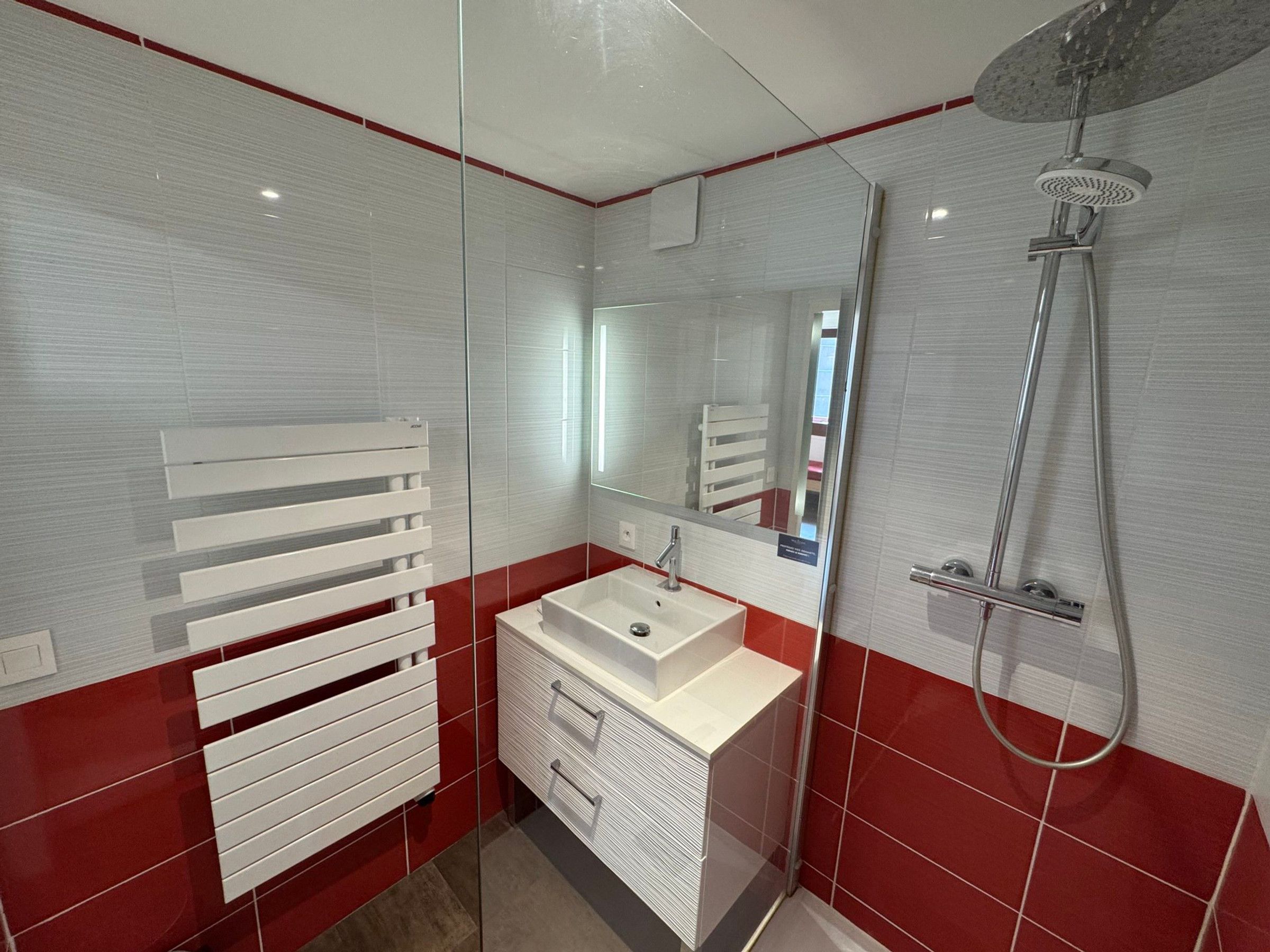 Een moderne badkamer met een douchecabine van glas, een wit badkamermeubel met lades en een rode wand betegeld met tegels.