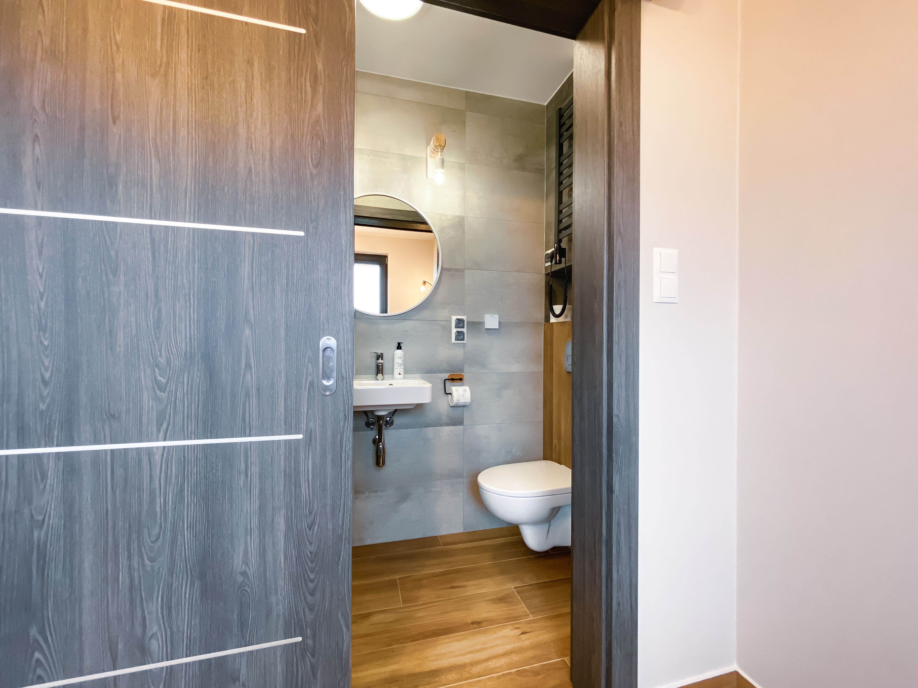 Un baño moderno con puerta de madera, espejo redondo, lavabo blanco y retrete.