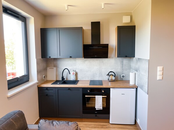 A compact kitchen with dark cabinets, a built-in oven, a dishwasher, and a refrigerator. - Pod Czwórką (Kosakowo)