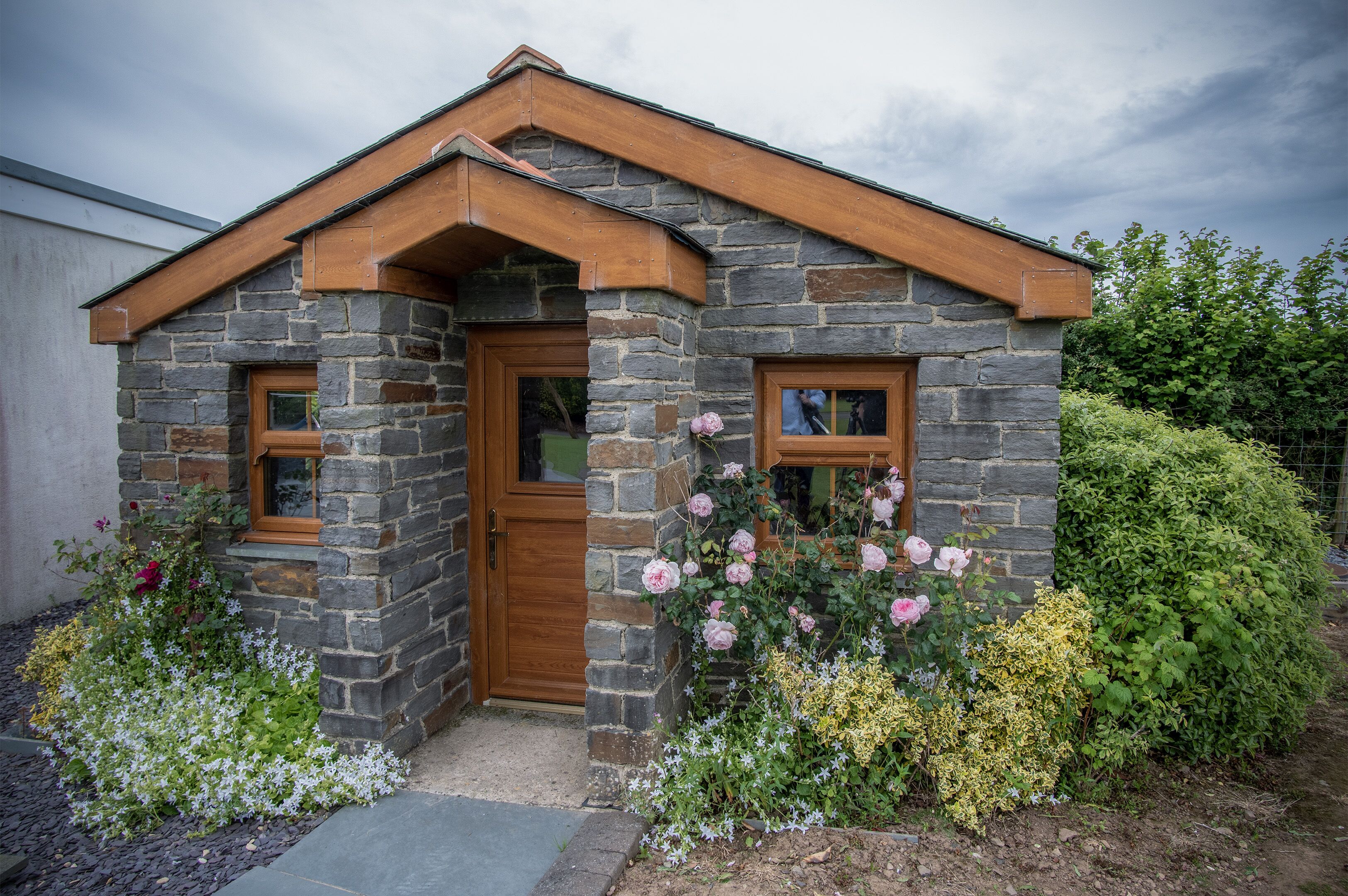 A stone house with a wooden door and windows, surrounded by various plants and flowers.