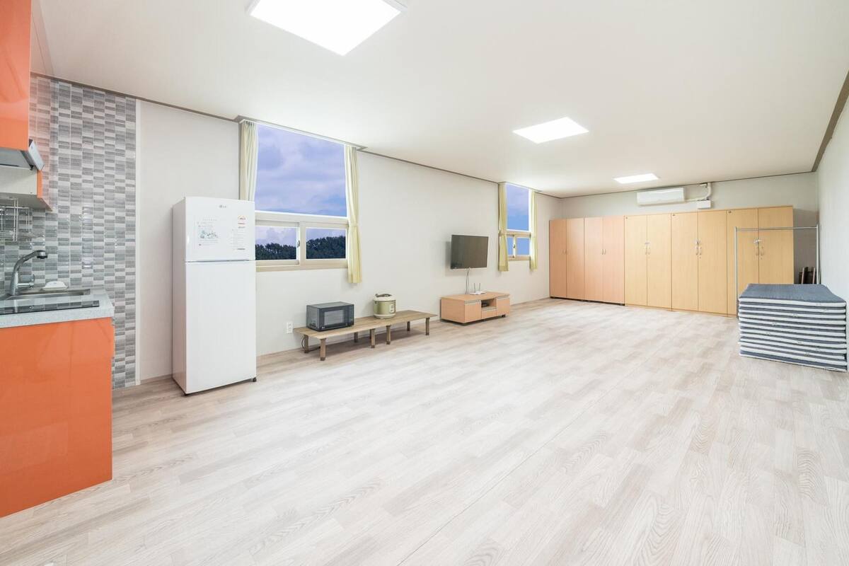 A spacious kitchen with a refrigerator, microwave, and sink. There is a television on a small table, and a large wardrobe in the background.