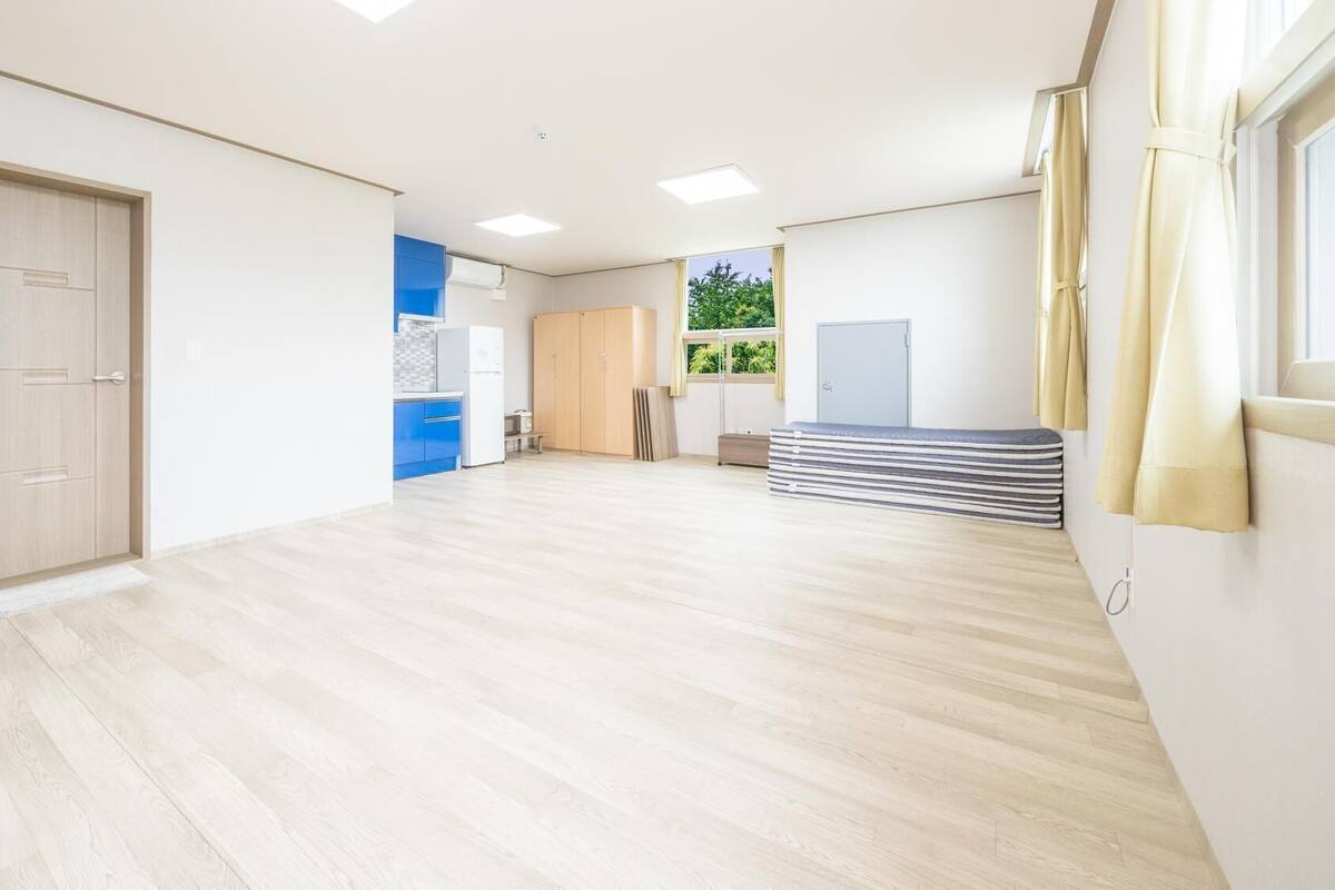 A spacious room with a wooden floor, a kitchenette, a wardrobe, and a window with curtains.