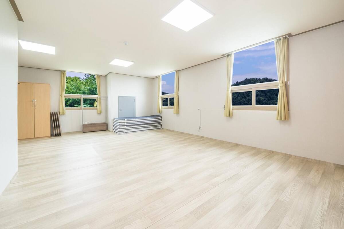 A spacious room with wooden flooring, large windows with curtains, a door, and a stack of mattresses.
