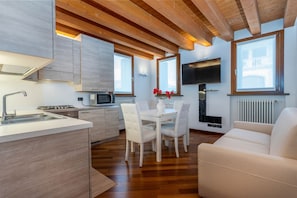 A modern kitchen with wooden beams, a dining area with white chairs, a TV mounted on the wall, and a sofa.