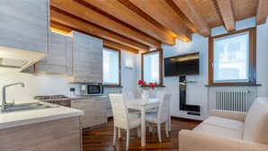 Una cocina moderna con vigas de madera, una zona de comedor con sillas blancas, un televisor montado en la pared y un sofá.