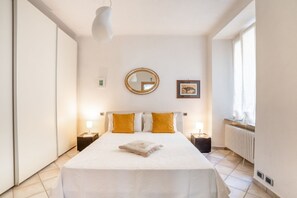 A bedroom with a bed, two bedside tables, a round mirror, a framed picture, and a window.