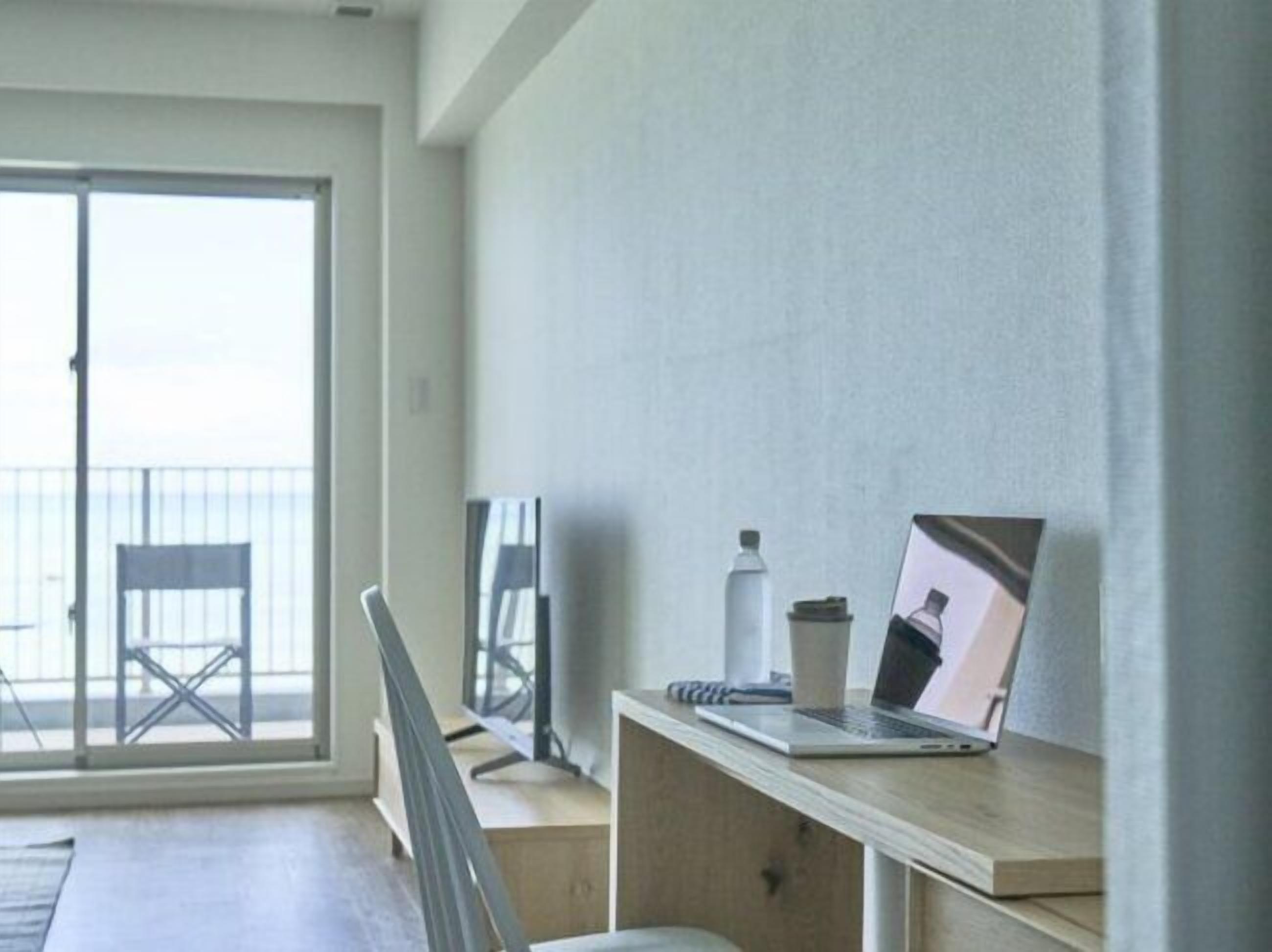 Et moderne værelse med en stor skydedør til en balkon, et træskrivebord med en bærbar computer, en hvid flaske og en notesbog.