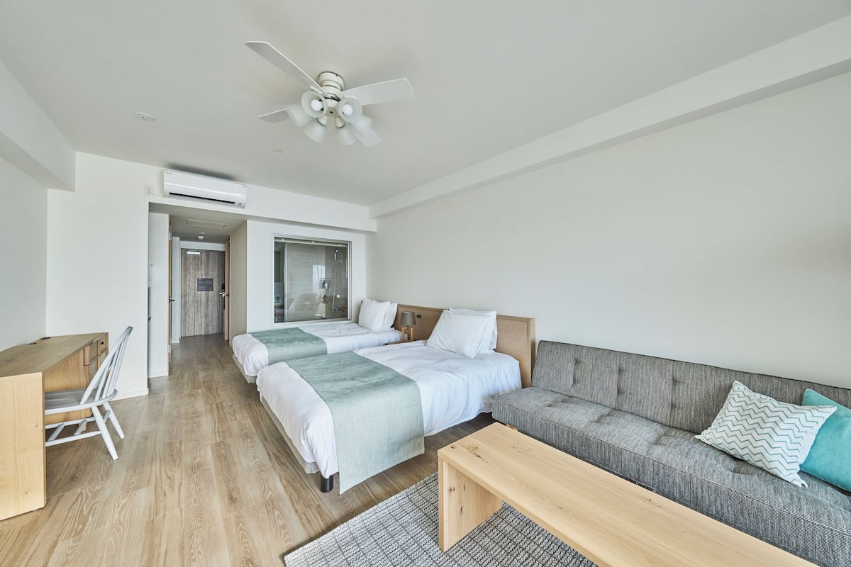 Ein modernes Hotelzimmer mit zwei Betten, einer Couch, einem Schreibtisch und einem Deckenventilator.
