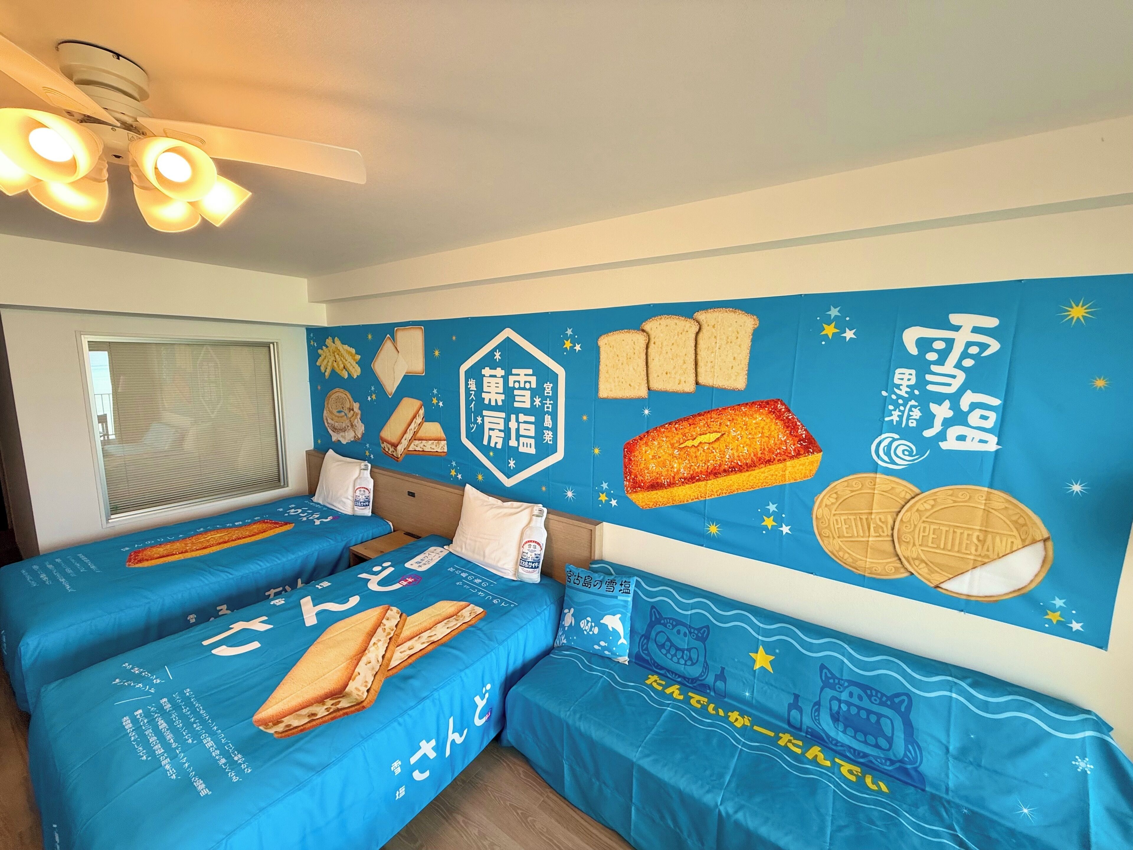 A hotel room with two beds, a ceiling fan, and a wall decorated with food illustrations and Japanese text.