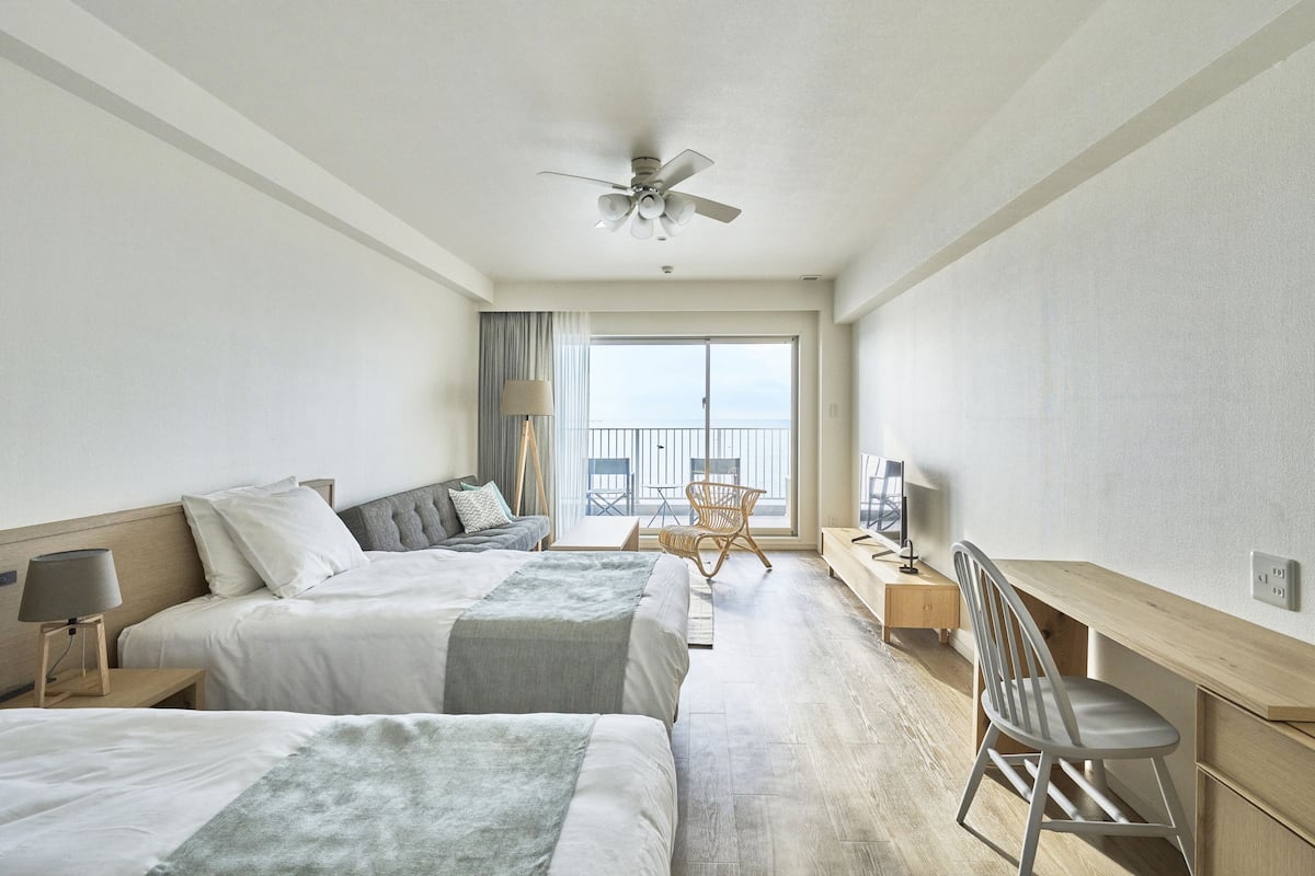 Ein Hotelzimmer mit zwei Betten, einem Schreibtisch, einem Sessel und einem Balkon, inklusive Deckenventilator.