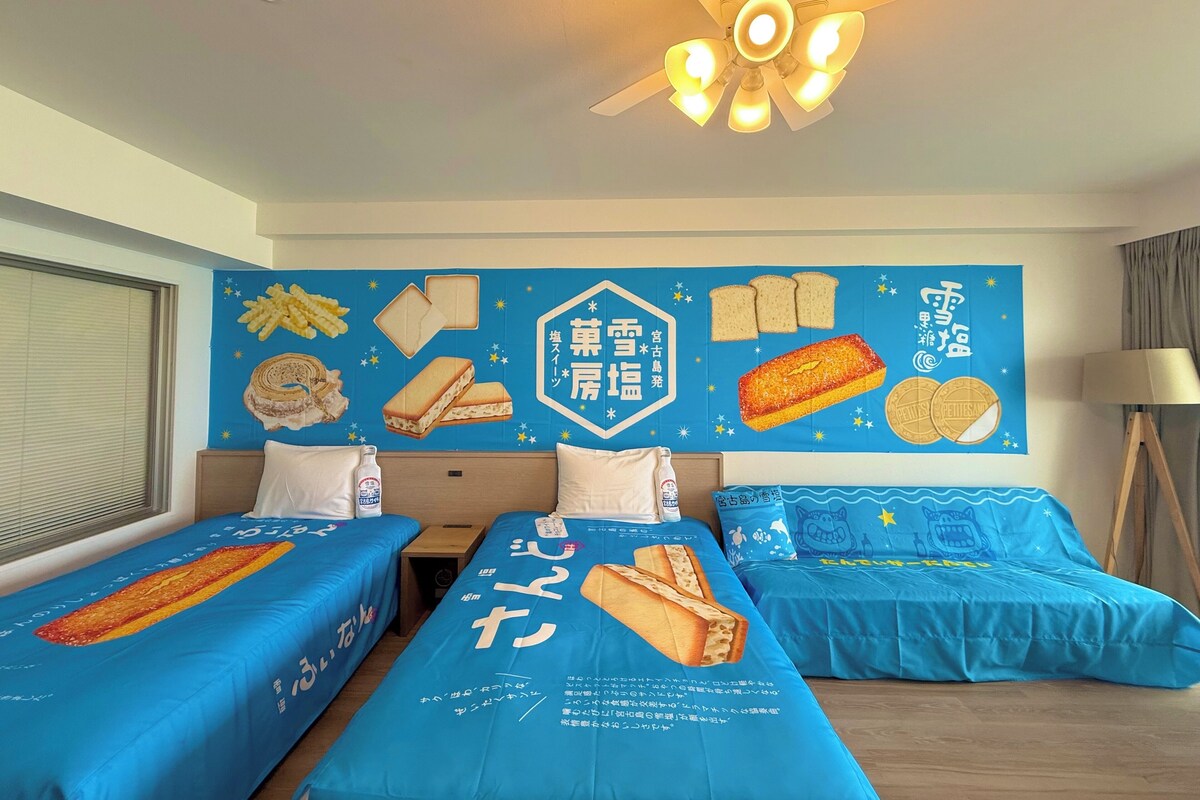 A hotel room with two beds, a ceiling fan, and a wall decorated with food items and Japanese text.