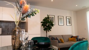Ein modernes Wohnzimmer mit einer Couch, einem Esstisch mit einer Vase und Gläsern, einer Küche im Hintergrund und drei gerahmten Bildern an der Wand.