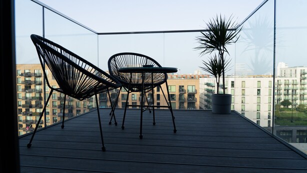 Ein Balkon mit zwei schwarzen Stuhlen aus Draht, einem Blumentopf und Blick auf Stadtgebäude.