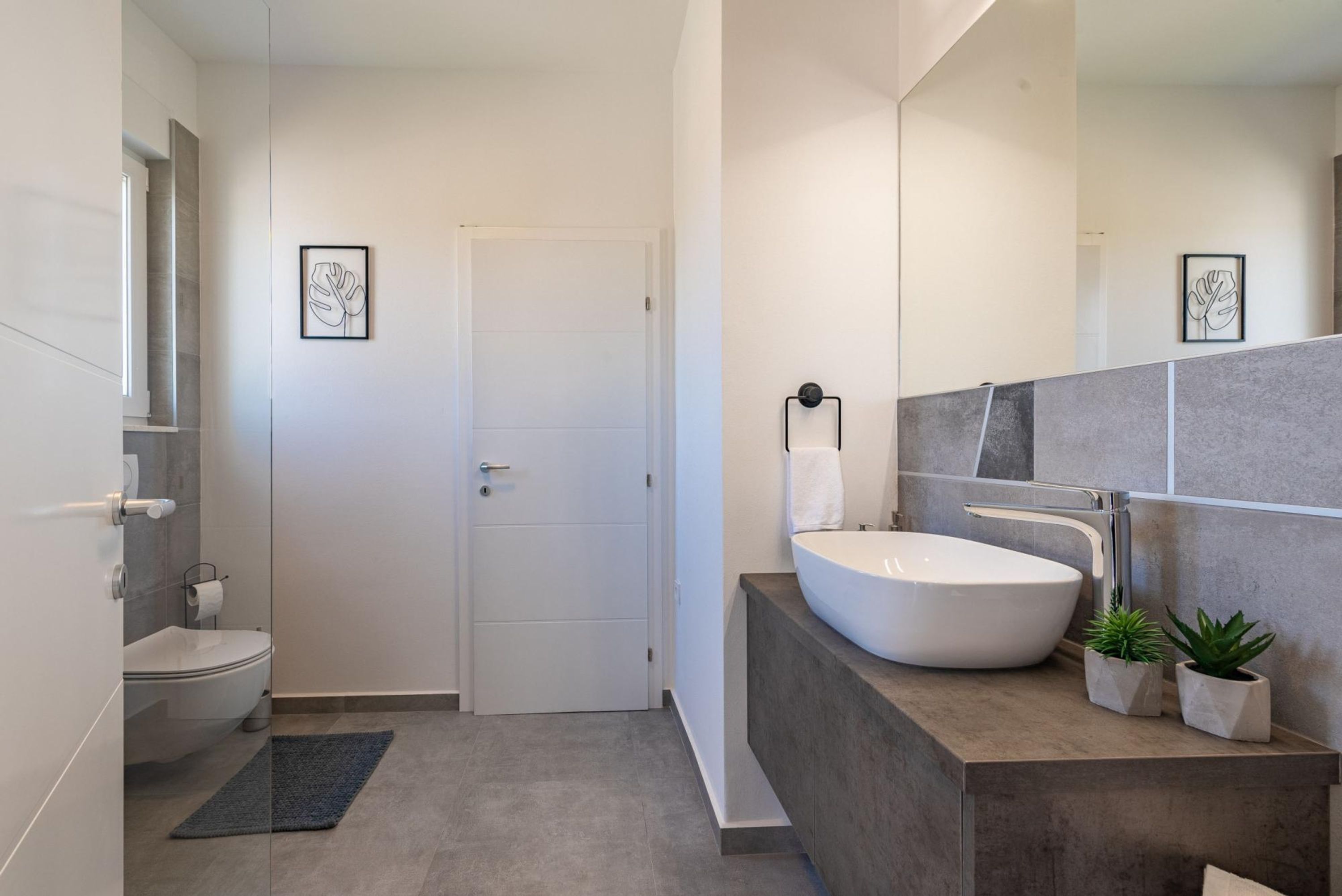 A modern bathroom with a white toilet, a large mirror, a white sink, and two small potted plants.