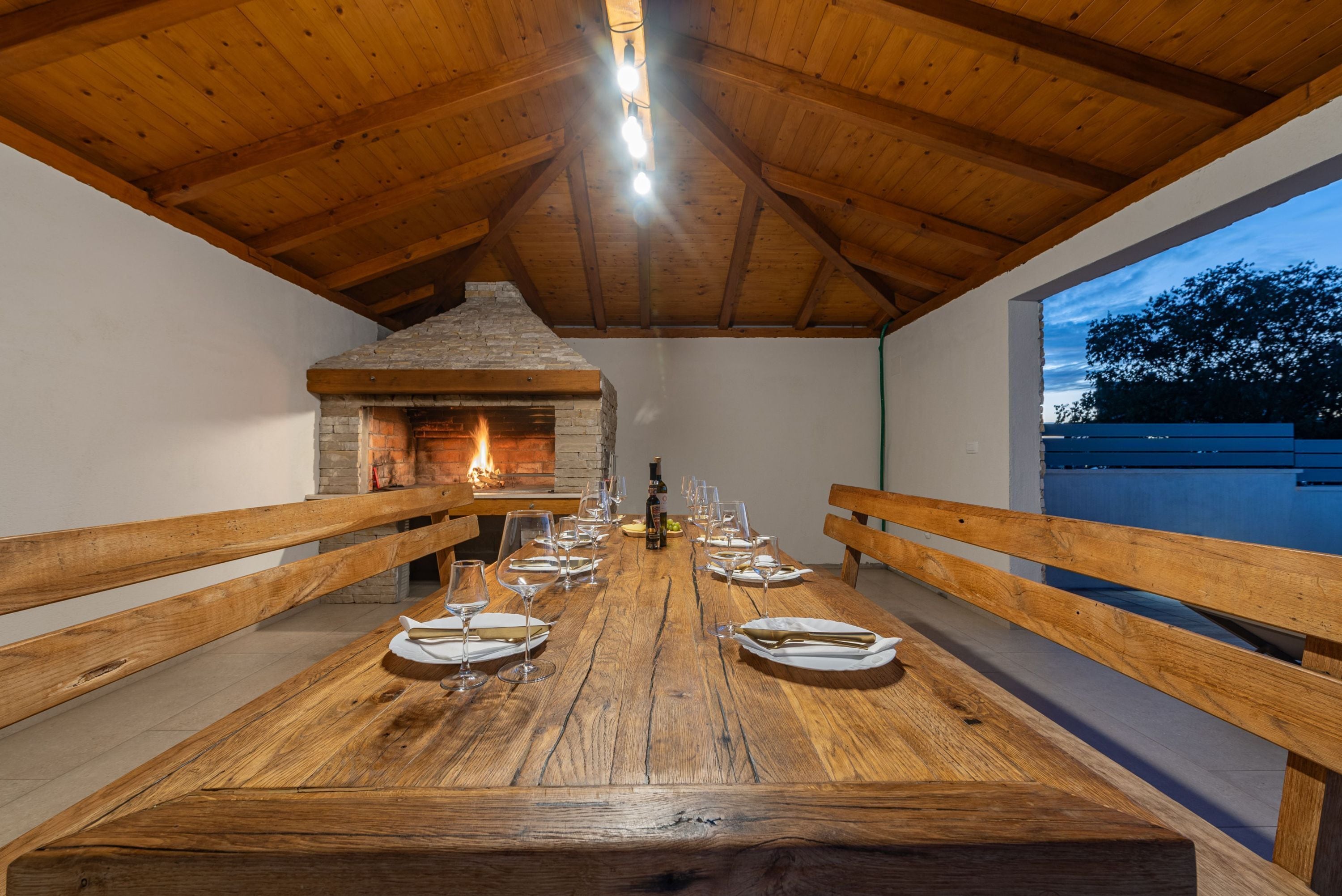Un lungo tavolo in legno apparecchiato per la cena, con panche su entrambi i lati, un camino in pietra sullo sfondo e una vista del cielo attraverso una finestra.