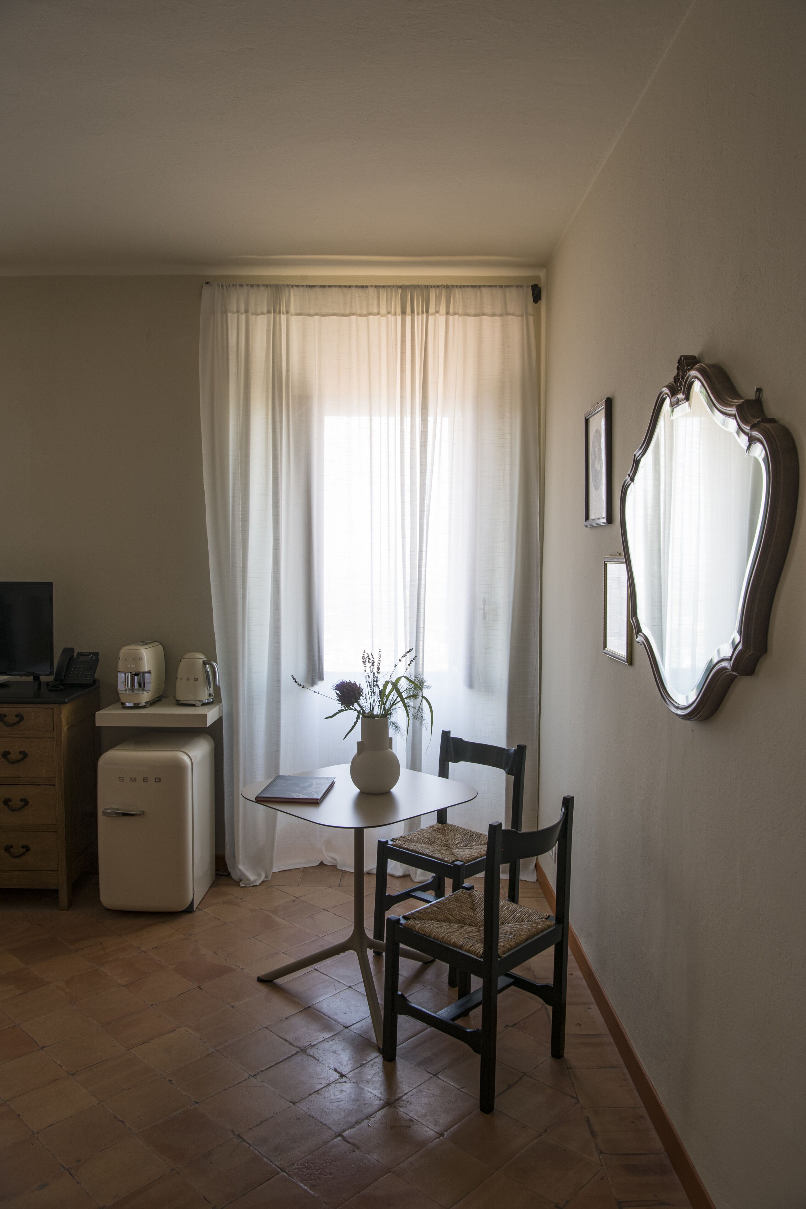 A small, well-lit room with a dining table, chairs, a small refrigerator, a television, and a window with curtains.