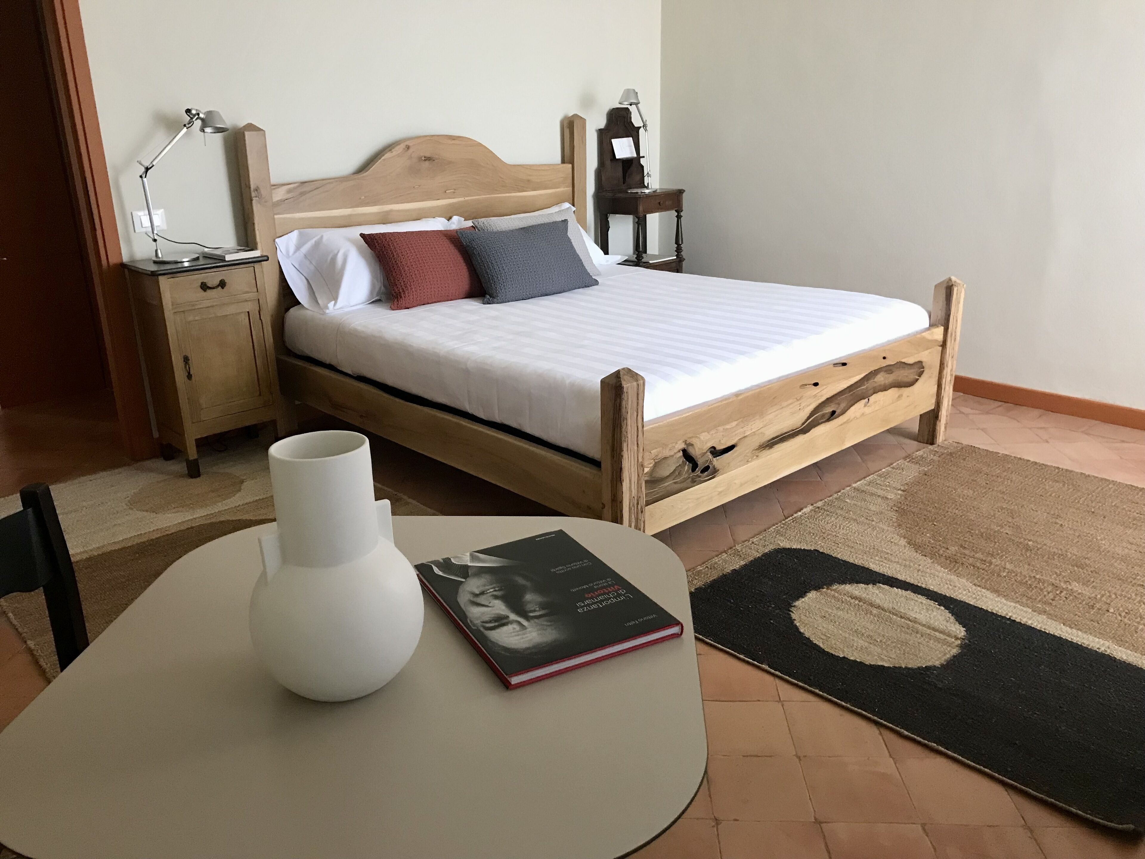 Une chambre à coucher bien rangée, avec un lit en bois, des lampes de chevet, une table de chevet sur laquelle repose un livre, un vase blanc et un tapis.
