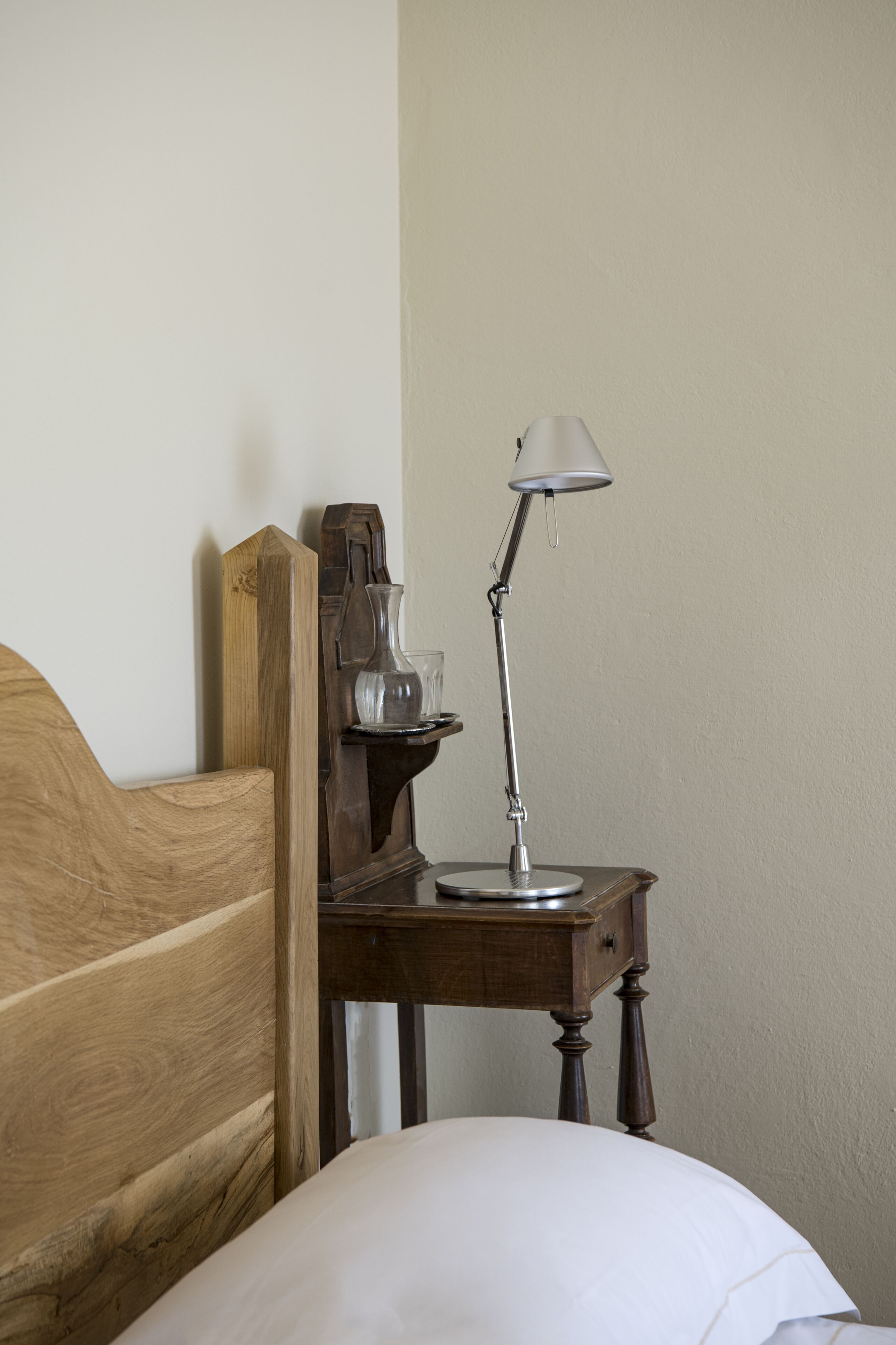 Ein hölzernes Bett mit einem weißen Kopfkissen, ein Nachttisch aus Holz mit einer Lampe und eine Vase darauf.