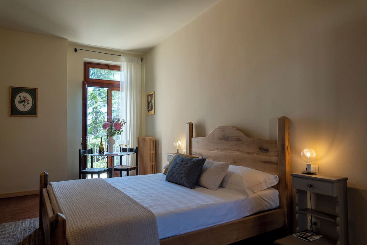 Een slaapkamer met een houten bed, nachtkastjes met lampen, een raam met gordijnen en een tafeltje met een vaas en bloemen.