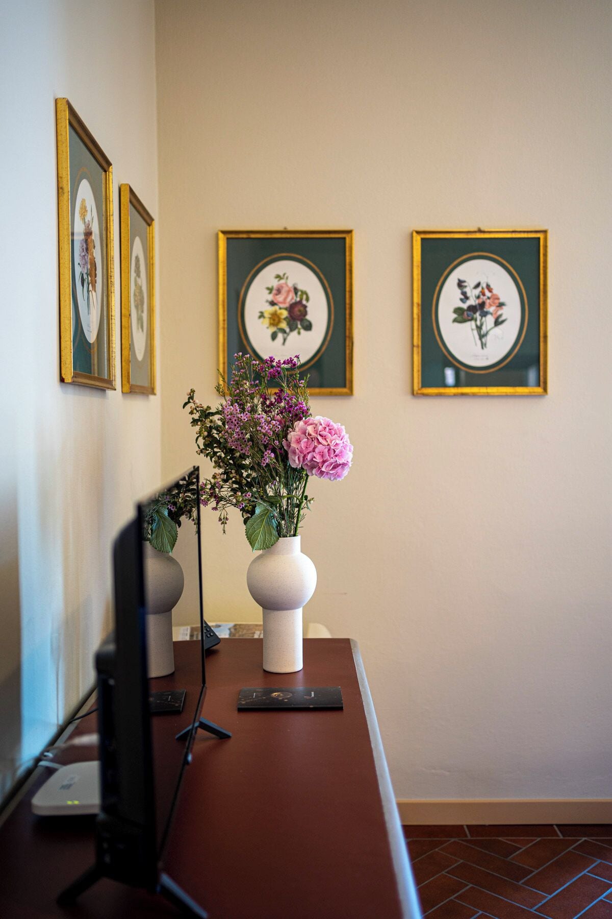 A desk with a vase of flowers, a computer monitor, and framed botanical prints on the wall.