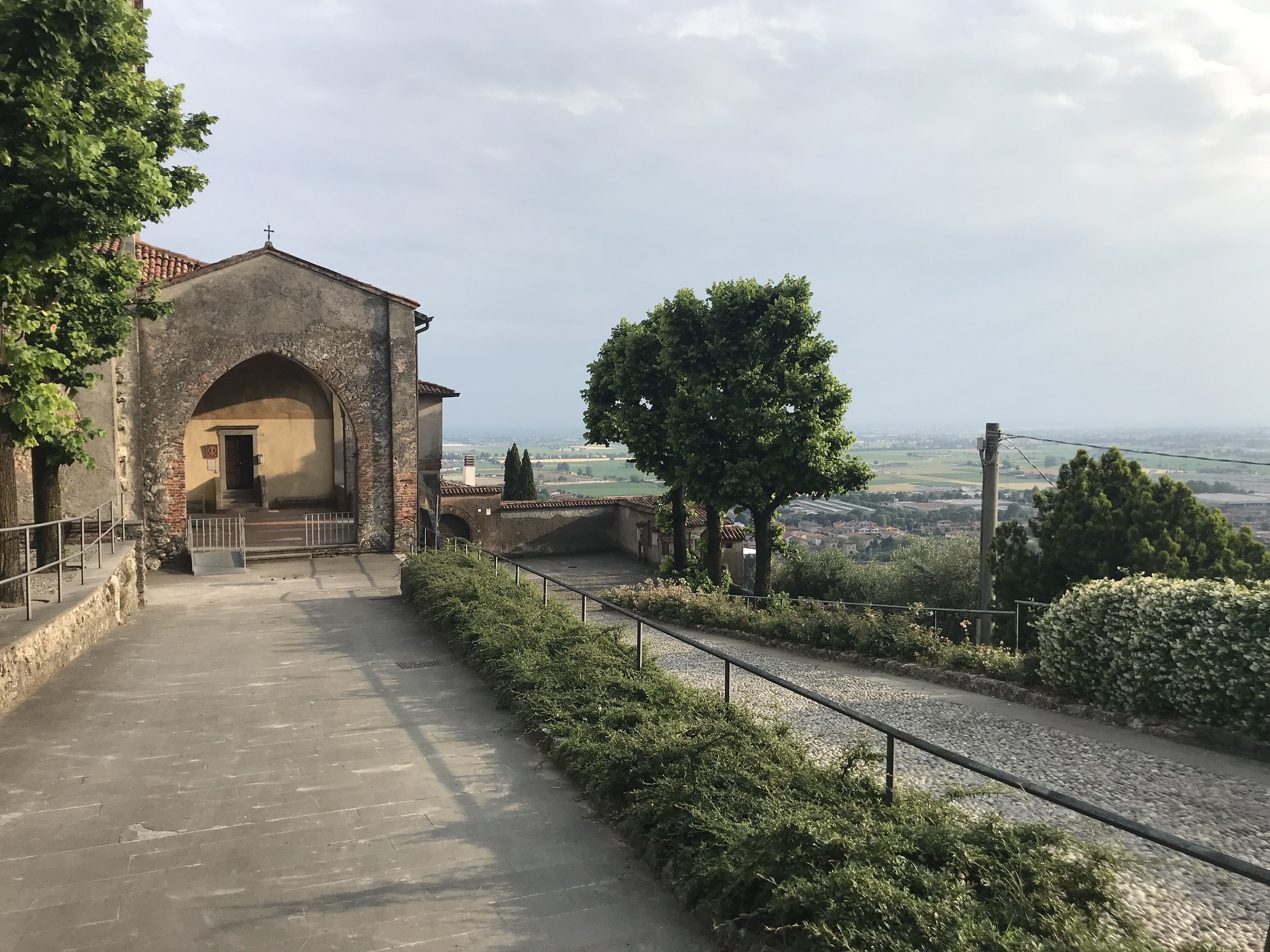 Un sendero conduce a un edificio histórico con una entrada en arco, rodeado de follaje y con vistas a un paisaje lejano.