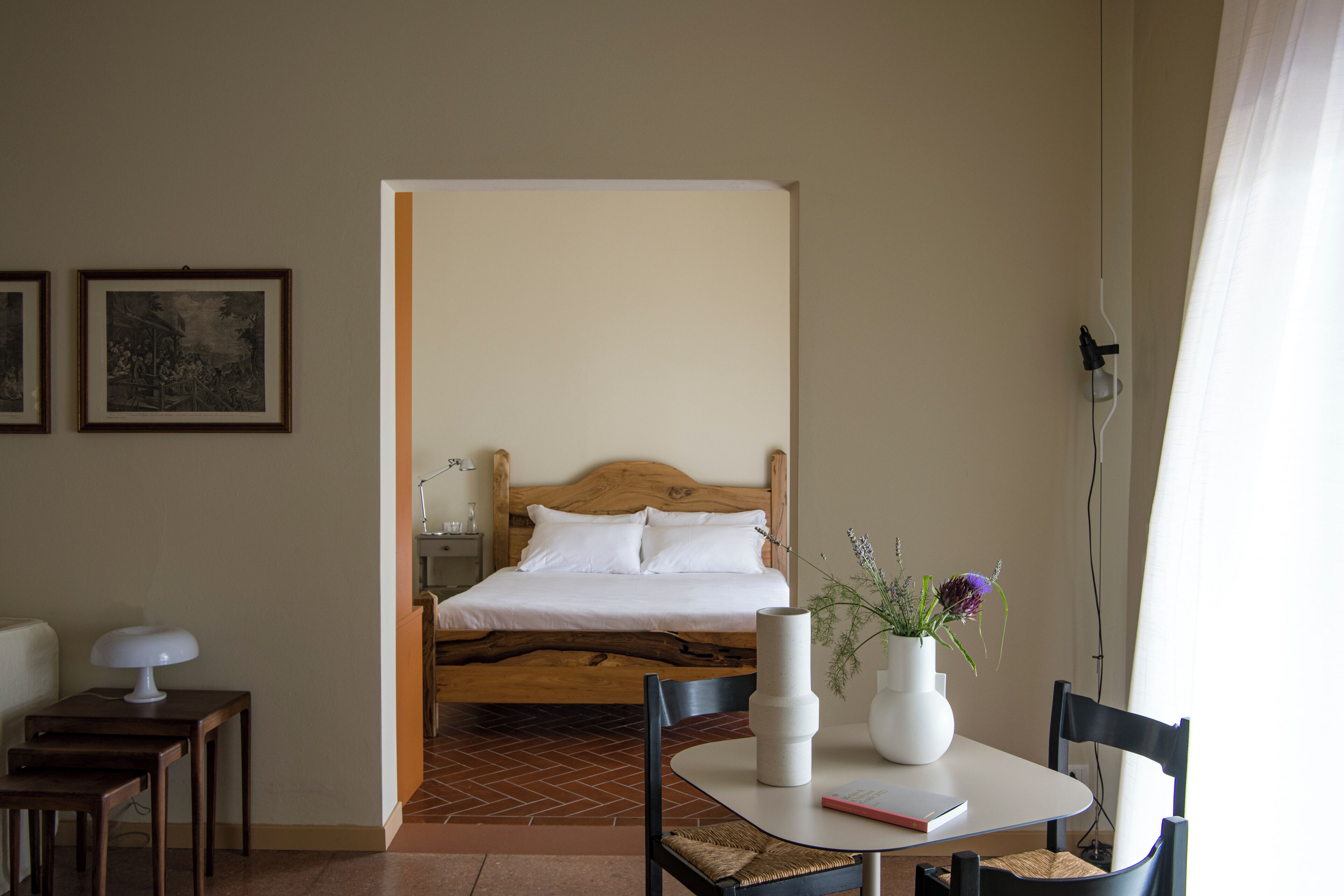 Une chambre à coucher avec un lit en bois, une table de chevet avec une lampe, une chaise, une petite table sur laquelle est posé un vase de fleurs, et un tableau encadré accroché au mur.