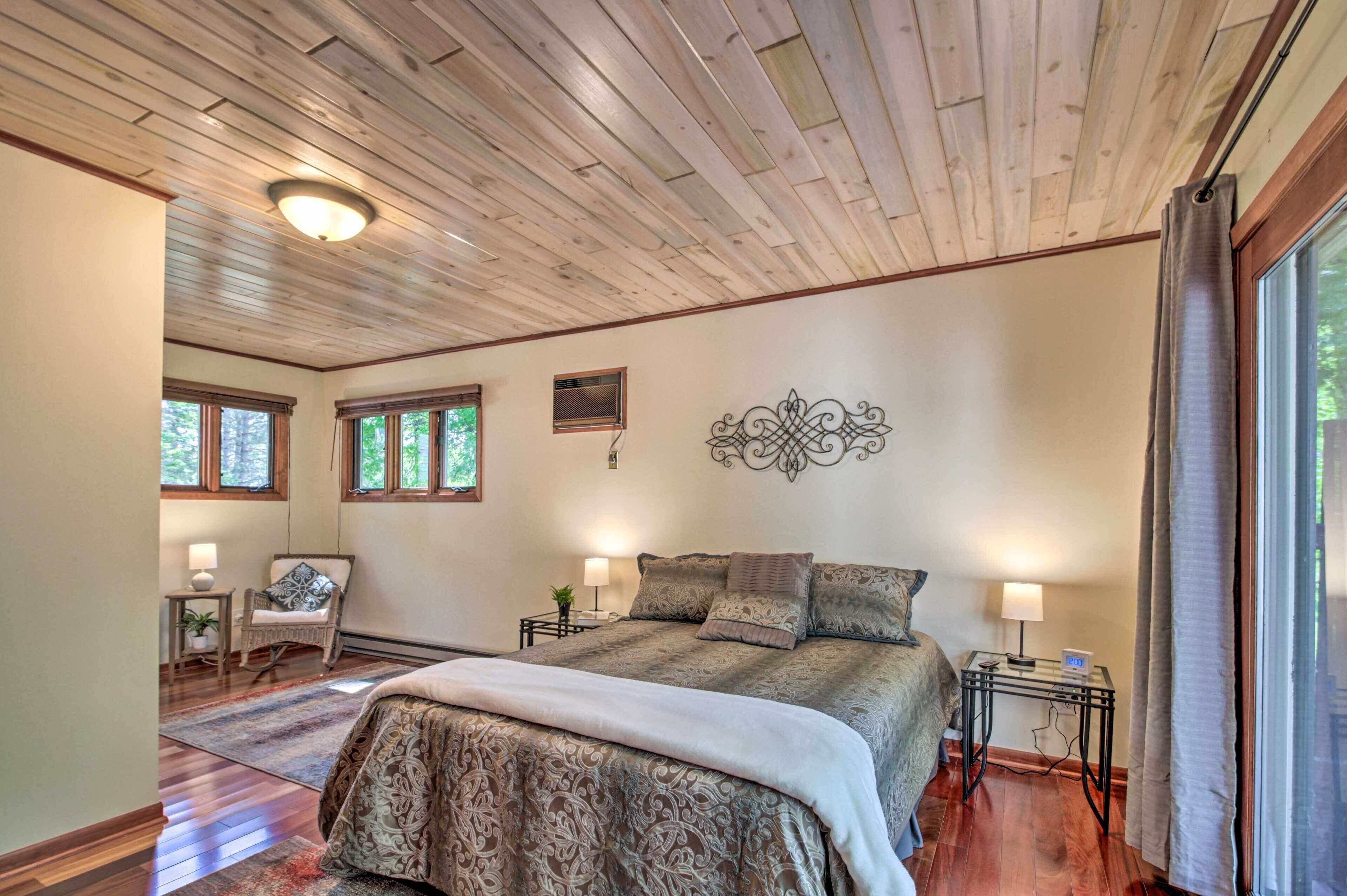A bedroom with a wooden ceiling, a large bed with patterned bedding, two bedside tables with lamps, a chair, and a decorative wall piece.