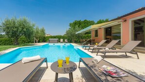 A swimming pool with lounge chairs and a table with drinks, surrounded by greenery and a building with large windows. - Pool, whirlpool, 12 persons, garden, children's playground,bicycles,table tennis (Anžici)