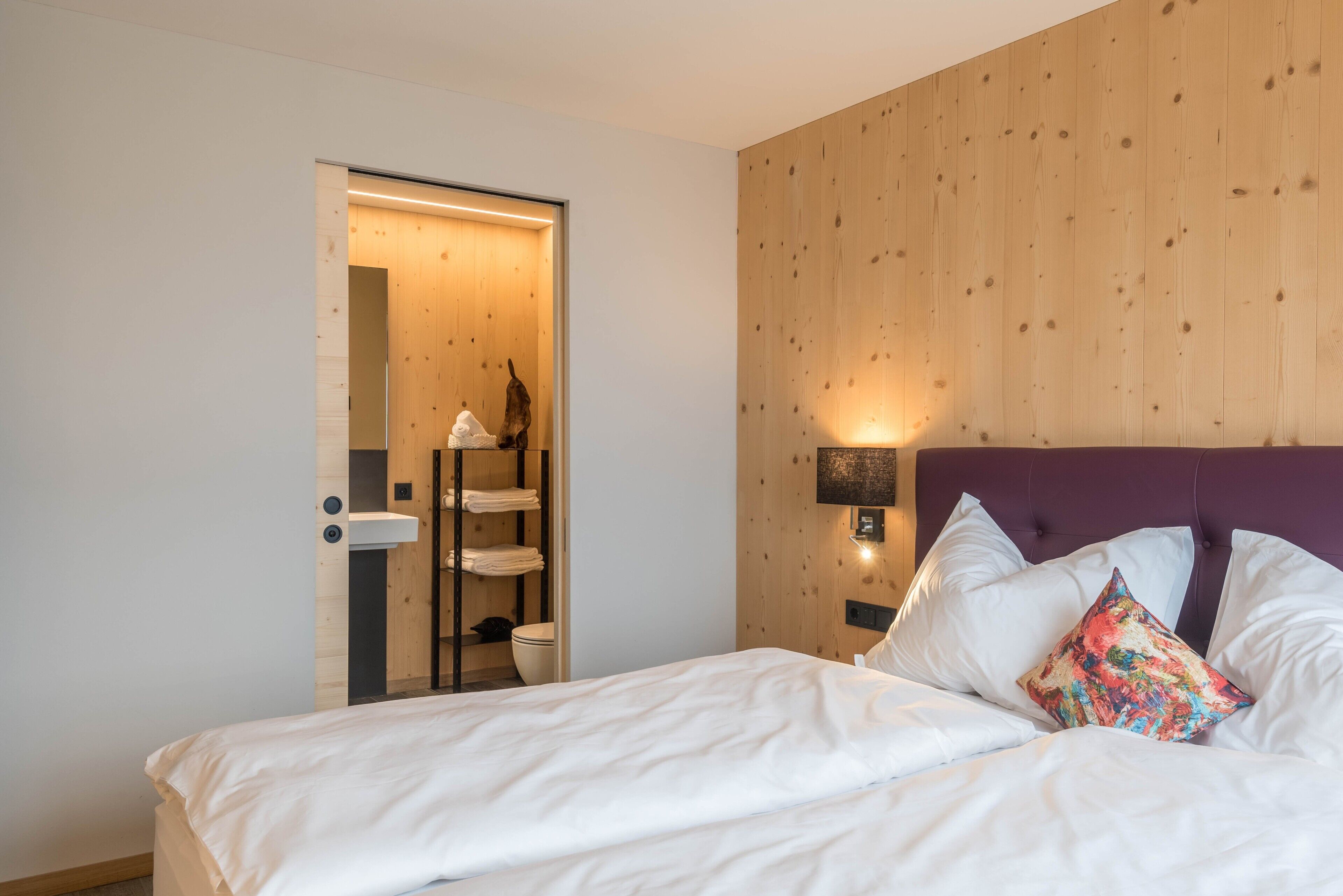 Ein Schlafzimmer mit Bett, Nachttisch, Wandleuchte, Wandverkleidung aus Holz und einem durchgehenden Türspalt sichtbaren Badezimmer.