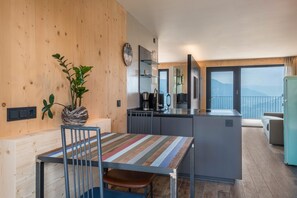 A modern kitchen with a central island, wooden walls, a dining area with a striped table, and a view of mountains through large windows. - 'AUMIA Apartment Turmalin' with Mountain View & Terrace (Völs)