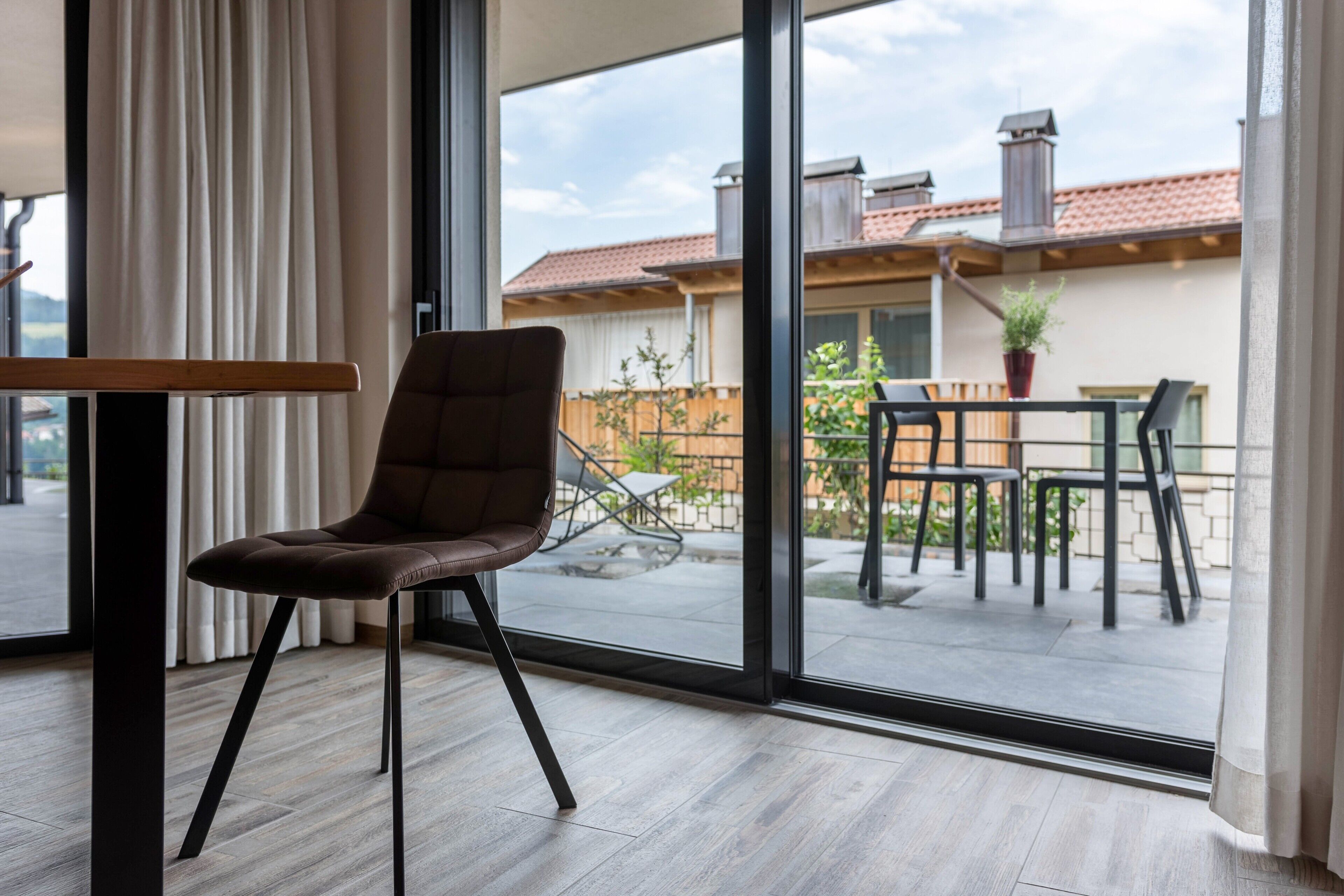 A modern room with a large window, a brown chair, a wooden table, and a view of a residential area.