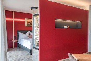 A modern hotel room with a red accent wall, a bed with white bedding, a bedside table, a mounted TV, and a stone wall feature.
