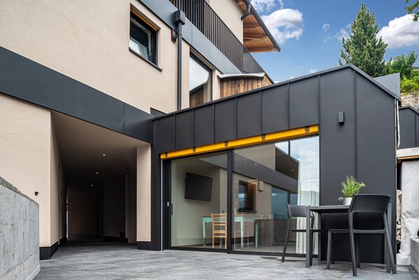 A modern building with a glass door entrance, a small outdoor seating area with chairs and a table, and a potted plant.