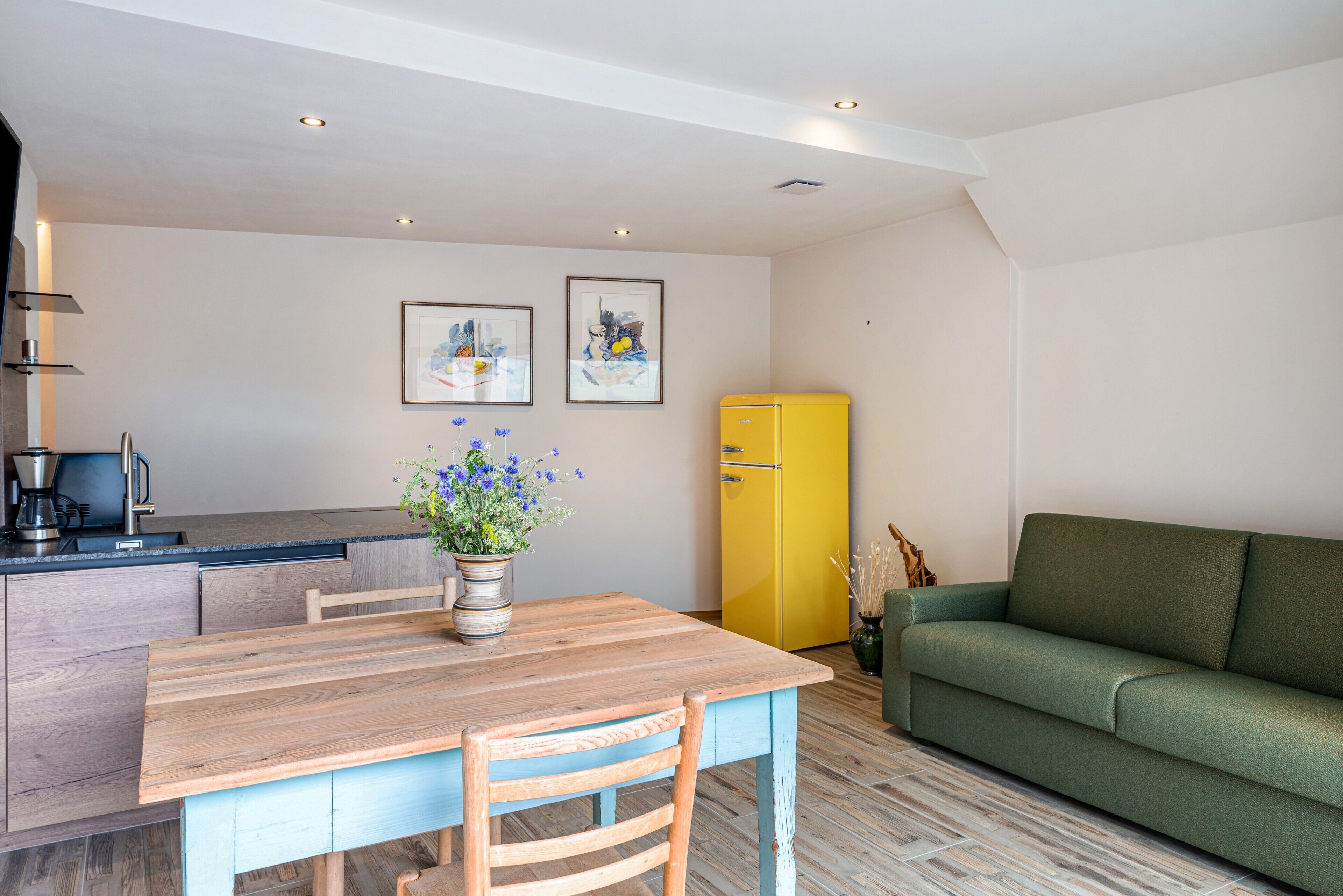 A compact living space with a wooden dining table, a green sofa, a yellow refrigerator, and a kitchen area with a coffee maker.