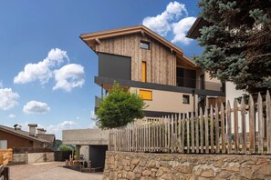 Un bâtiment moderne à deux étages, doté d’un mur en pierre et d’une clôture en bois, avec un balcon et un petit arbre.
