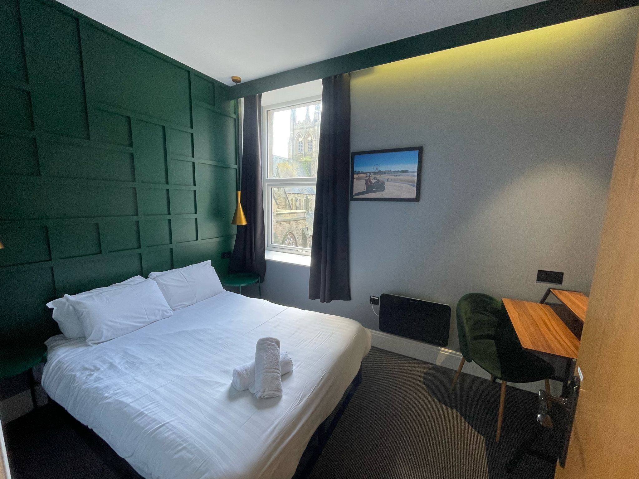 A hotel room with a large bed, a desk, and a chair. There is a window with curtains and a painting on the wall.