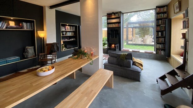 A modern living room with a wooden dining table, bookshelves, a fireplace, and a large window overlooking a garden.
