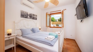 Slaapkamer met bed, nachtkastje, lamp, plafondventilator, televisie, raam en kunstwerk.
