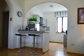 A kitchen with a bar stool, a clock, and a window with curtains.