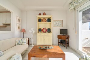 Un salon comprenant un canapé, une table basse en bois, un meuble avec des assiettes décoratives, un téléviseur et un ventilateur.